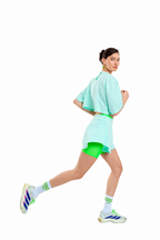 A woman in a mint green athletic set consisting of a short-sleeve crop top, matching shorts, and bright green fitted liner shorts, running against a solid white background. She is wearing white socks with green stripes and Adidas sneakers.