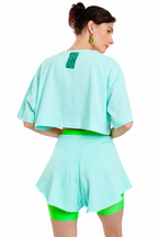 A model shown from the back wearing a mint green cropped t-shirt with a green patterned accent patch on the upper back, matching mint green shorts, and neon green leggings against a plain white background.