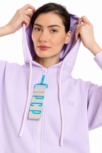 A woman wearing a light purple zip-up hoodie with white drawstrings and a gray zipper pull featuring bright blue accents. She is adjusting the hood of the sweatshirt against a plain white background.