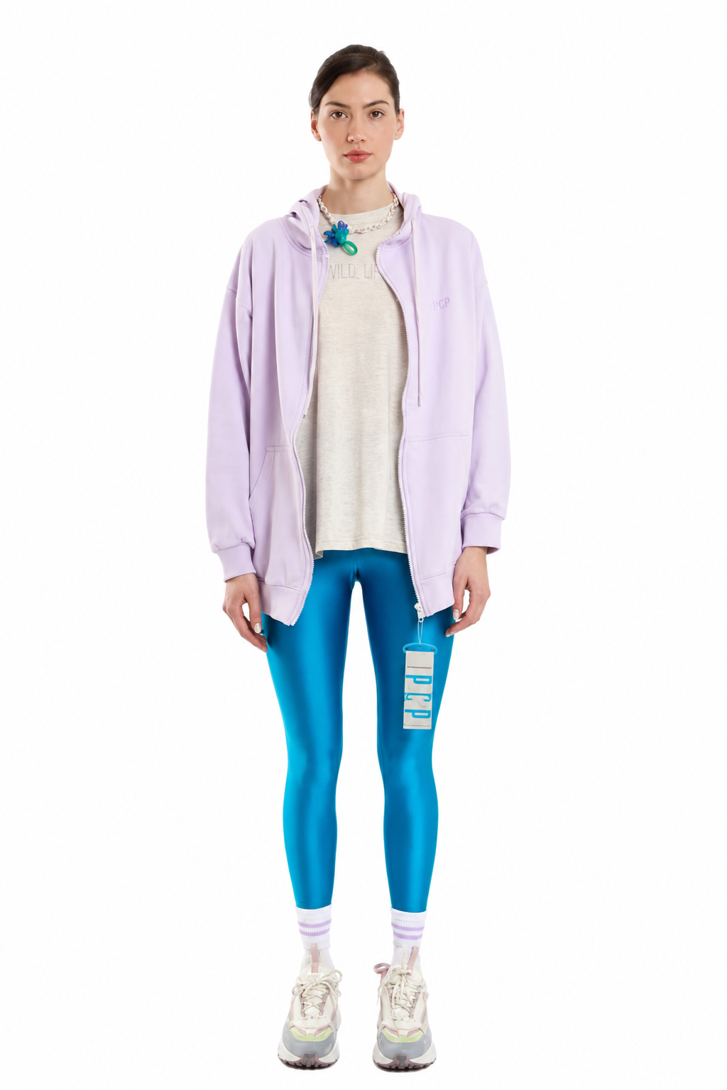 A model stands against a white background wearing a light purple zip-up hoodie over a cream knit top, bright blue leggings with a ripped detail on the left thigh, white socks, and light-colored sneakers.