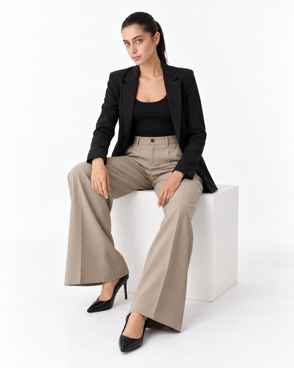A woman sits on a white cube in a studio setting, wearing a black blazer, black top, beige wide-leg pants, and black pointed heels.