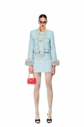 A model stands against a white background wearing a light blue tweed jacket and matching mini skirt with gray faux-fur trimmed cuffs, sheer beige lace tights, layered pearl necklaces, black high heels, and a small pink quilted handbag.