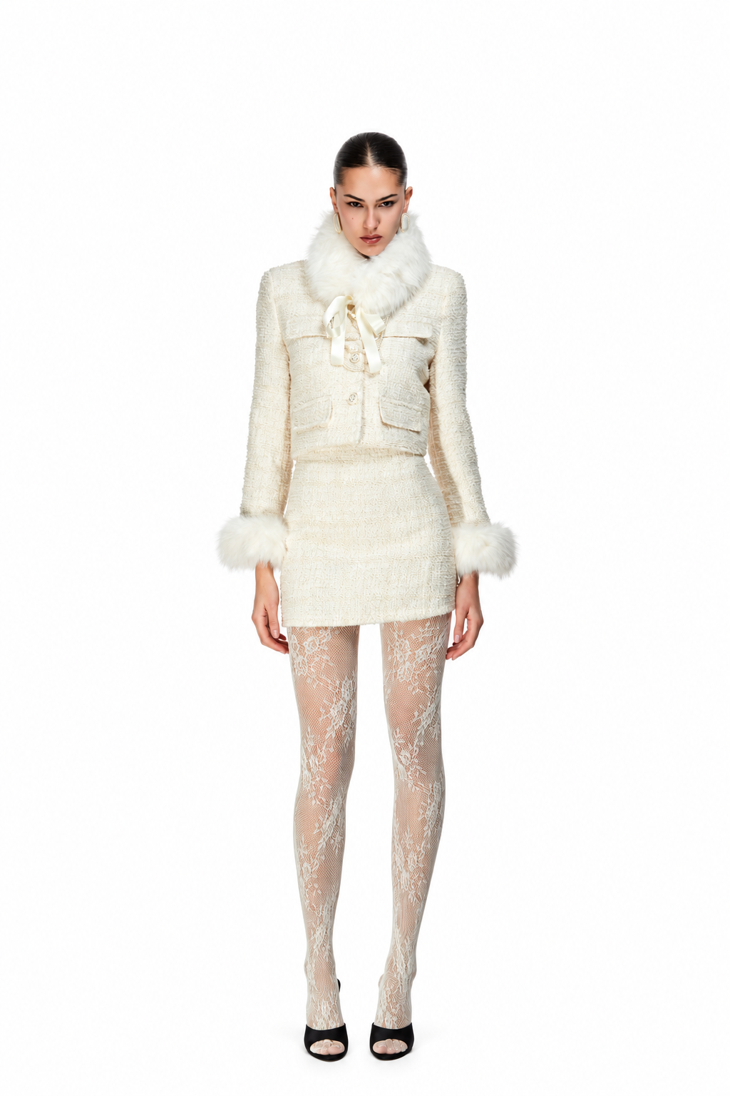 A model stands against a solid white background wearing a cream-colored tweed jacket and matching mini skirt set with faux fur trim, paired with sheer patterned tights and black heels.