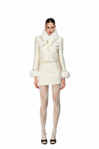 A model stands against a solid white background wearing a cream-colored tweed jacket and matching mini skirt set with faux fur trim, paired with sheer patterned tights and black heels.