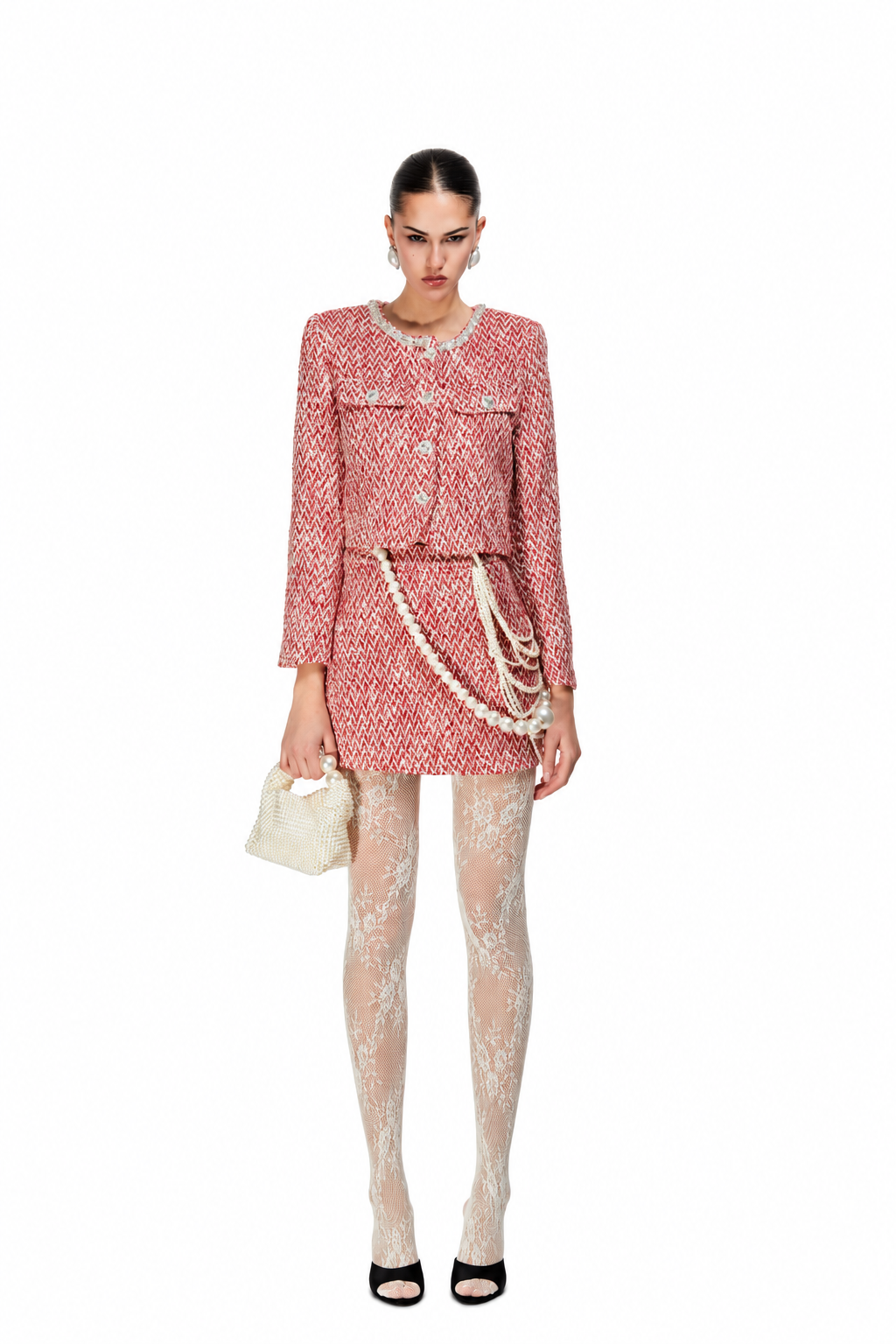 A model stands against a white background wearing a pink and white tweed mini dress with a matching jacket, patterned sheer tights, a small cream handbag, and black heels.