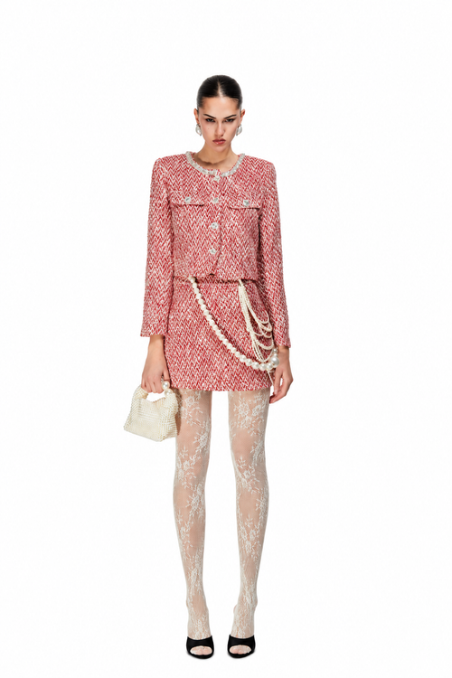 A model stands against a white background wearing a pink and white tweed mini dress with a matching jacket, patterned sheer tights, a small cream handbag, and black heels.