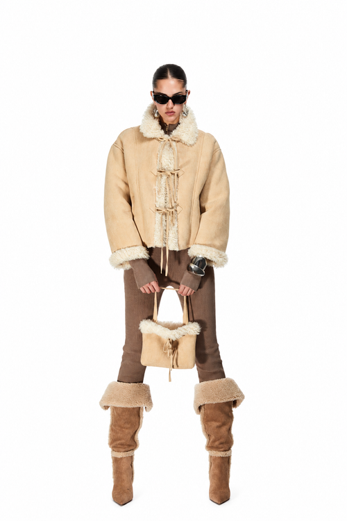 A model stands against a white background wearing a beige shearling-lined jacket with toggle closures, brown pants, tall brown boots with shearling trim, and holding a matching beige shearling handbag.