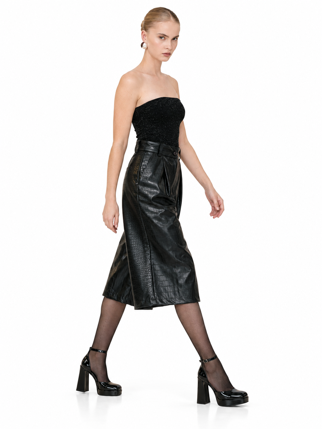 A model wearing a black strapless top and a black faux-leather midi skirt with a matching belt, styled with sheer black tights and black platform heels, against a plain white background.