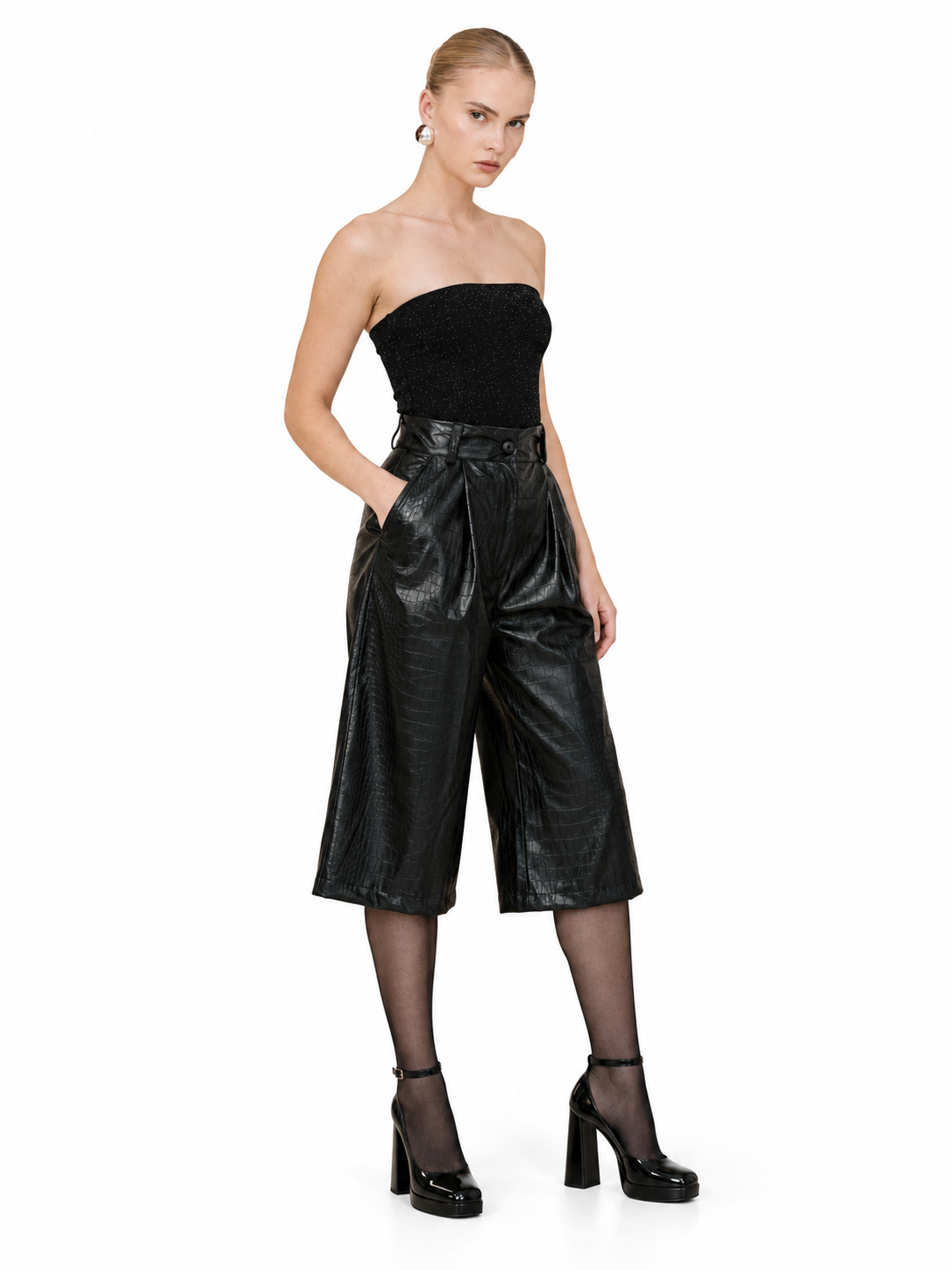 A model wearing a black strapless tube top and shiny black faux-leather cropped culottes, paired with sheer black tights and black platform heels, against a plain white background.