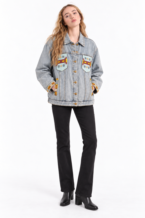A woman modeling a light-wash denim jacket with colorful embroidered patches on the chest and cuffs, paired with black pants and black boots, against a plain white studio background.