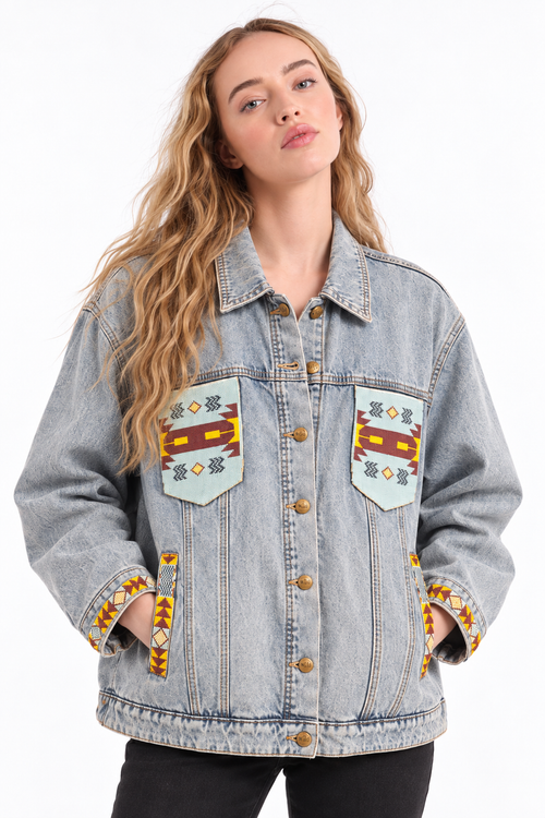 A woman wearing a light-wash denim jacket with colorful geometric embroidered patches on the chest and cuffs, photographed against a plain white background.