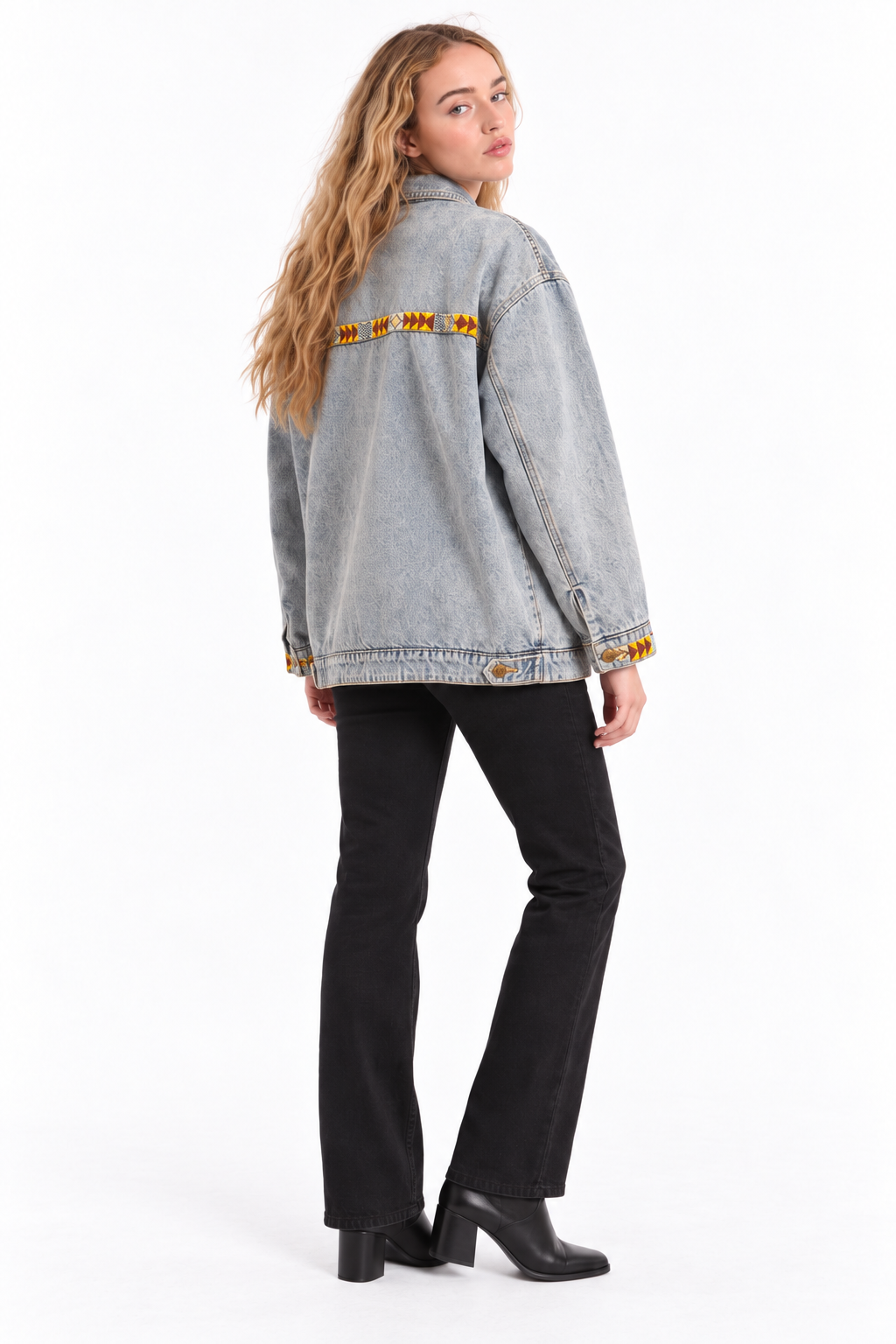 A woman modeling a light gray washed denim jacket with yellow embroidered details on the shoulders and cuffs, paired with black pants and black boots, photographed against a plain white studio background.
