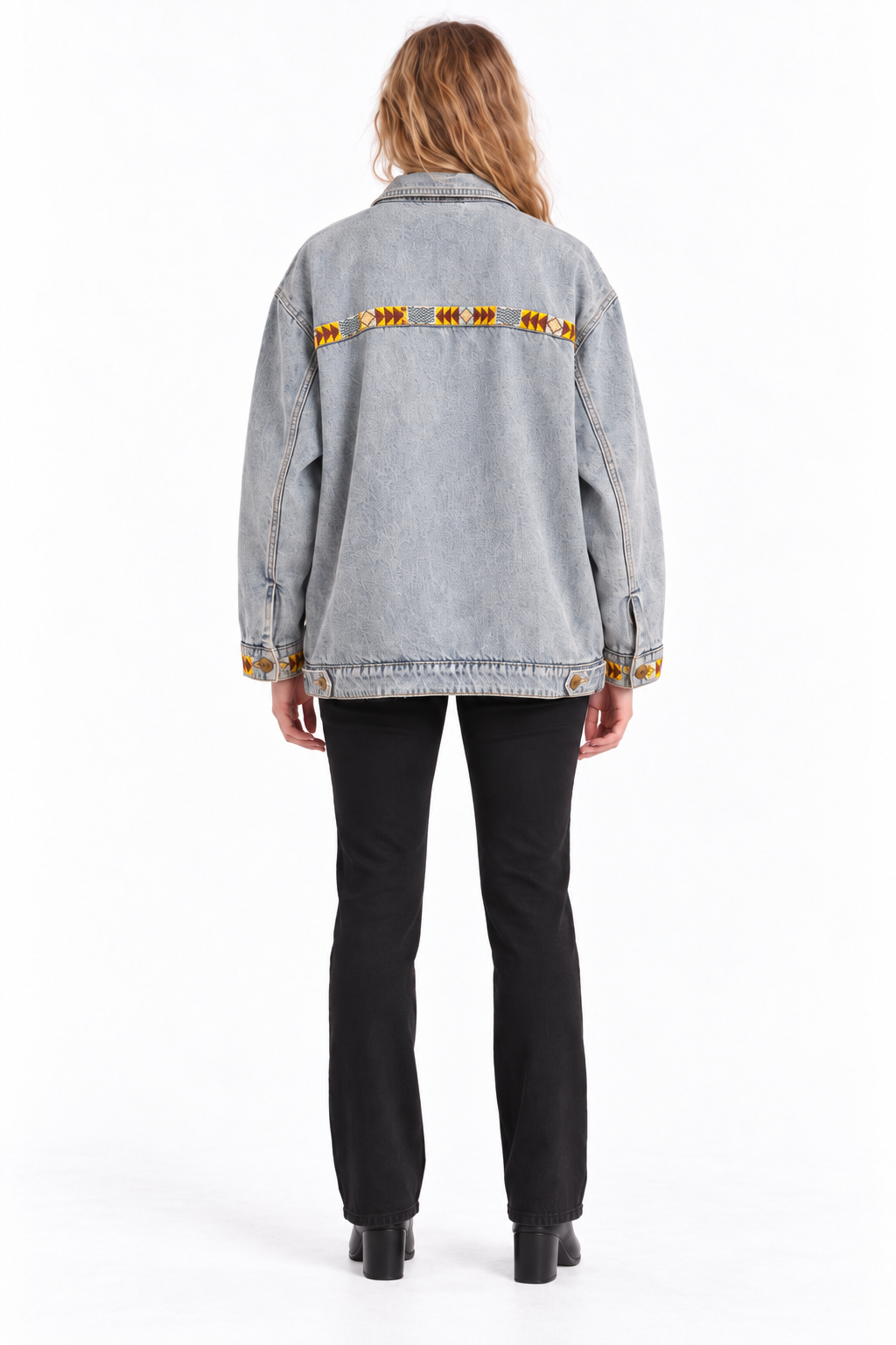 A model viewed from the back wearing a light gray denim jacket with a horizontal row of orange and yellow embroidered details across the upper back, paired with black pants and black boots, on a white studio background.