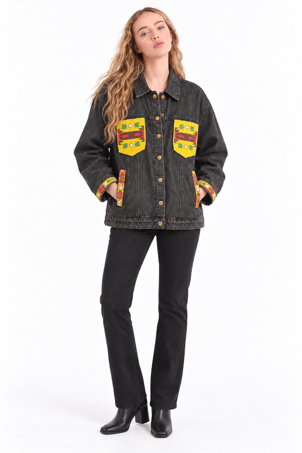 A woman modeling a dark washed denim jacket with colorful geometric patterned chest pockets and matching patterned pocket trim, paired with black pants and black boots against a plain white studio background.