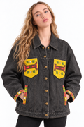 A woman wearing a dark gray washed denim jacket with yellow, red, and green geometric patterned chest and pocket patches, shown against a plain white background.