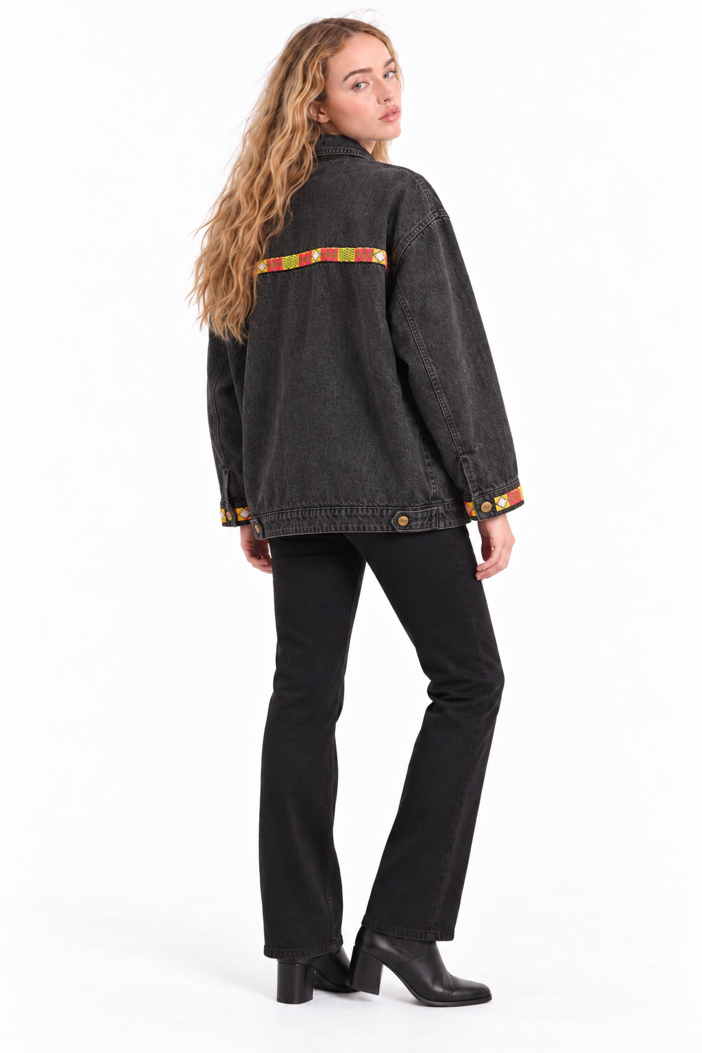 A woman modeling a washed black denim jacket with a colorful striped accent across the back and matching striped trim on the cuffs, paired with black pants and black boots, shot against a plain white studio background.