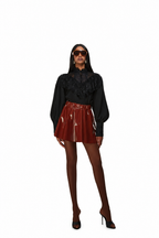 A model wearing a black long-sleeve blouse with ruffles and a shiny red mini skirt, paired with brown tights and black high heels, set against a plain white background.