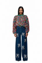 A model stands against a white background wearing a vibrant, multicolored fuzzy long-sleeve top and dark blue wide-leg jeans embellished with white bow patterns.