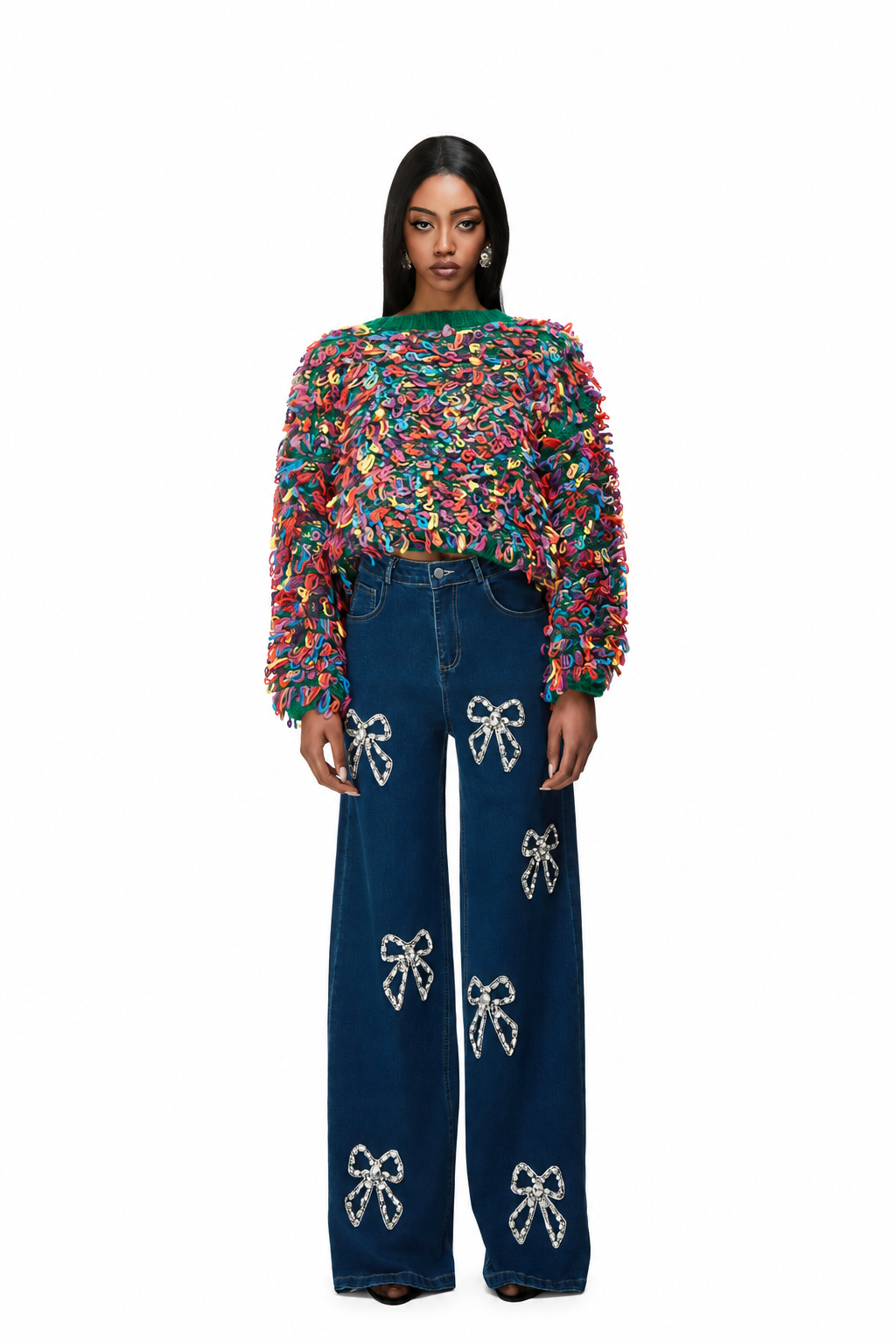 A model stands against a white background wearing a vibrant, multicolored fuzzy long-sleeve top and dark blue wide-leg jeans embellished with white bow patterns.