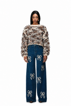 A model stands against a white background wearing a brown and beige textured fuzzy cropped sweater and dark blue wide-leg jeans decorated with white bow embellishments.