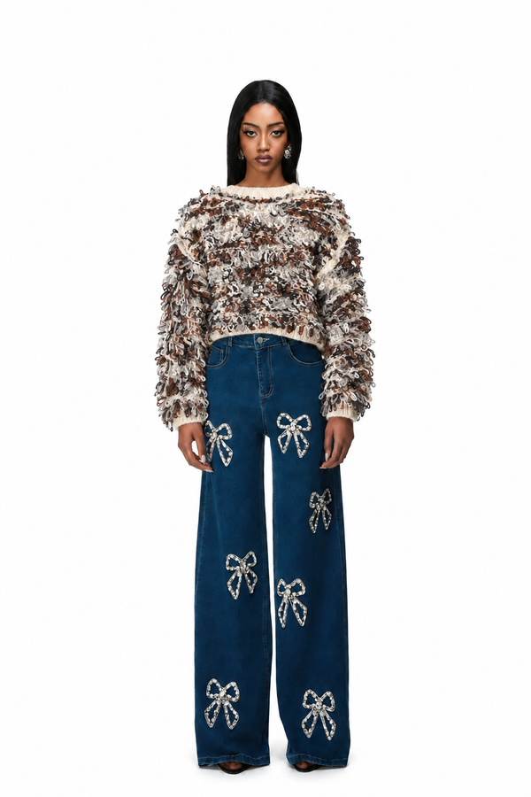 A model stands against a white background wearing a brown and beige textured fuzzy cropped sweater and dark blue wide-leg jeans decorated with white bow embellishments.