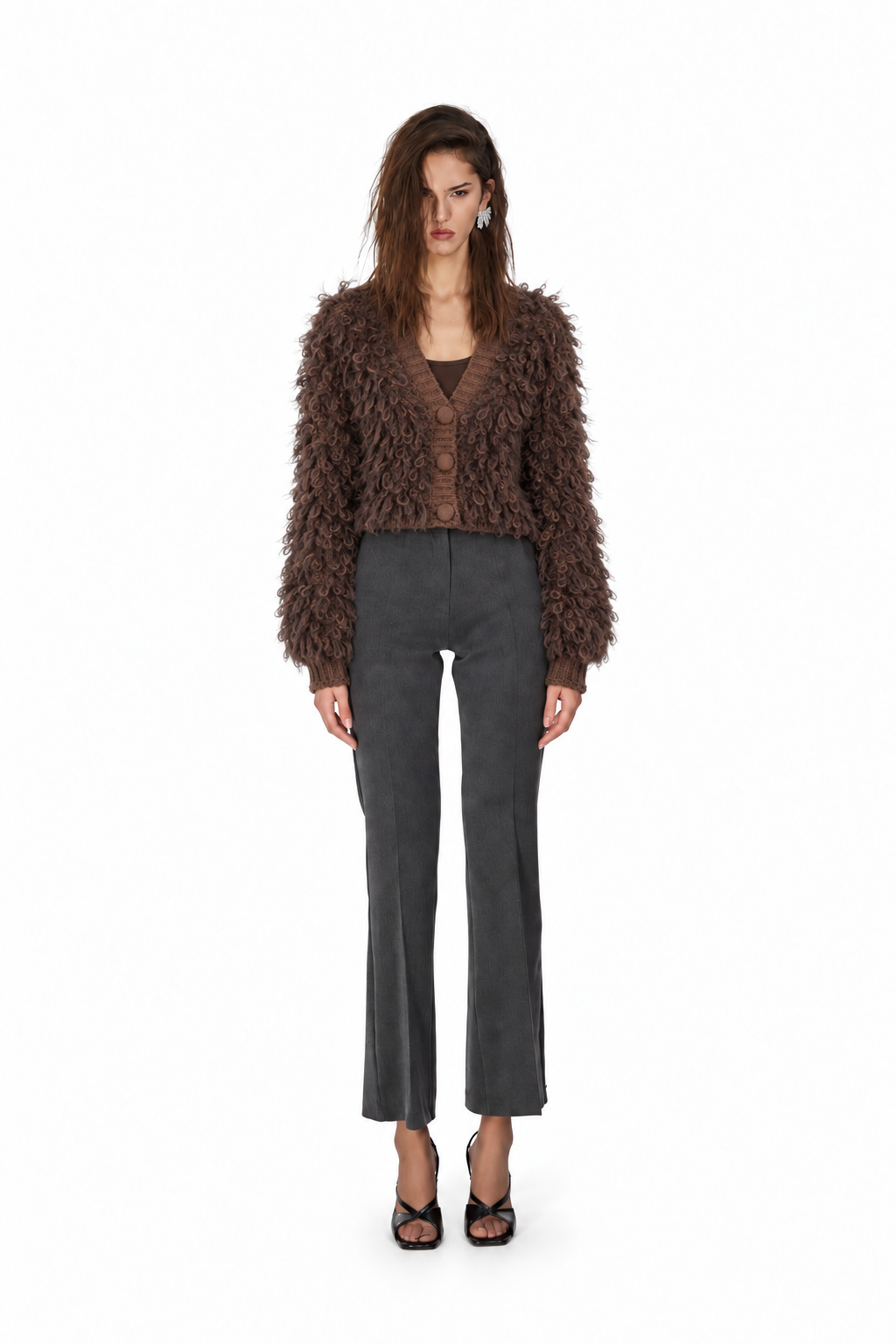 A model stands against a plain white background wearing a brown shaggy cropped cardigan, dark gray cropped flare pants, and black strappy heels.