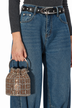 A person wearing blue jeans, a black star-spangled belt, and a dark ribbed top holds a small rhinestone bucket bag with a metallic gold handle against a plain white background.