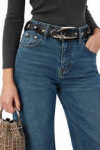 A close-up of a model wearing blue jeans with a black belt featuring silver star details and a large silver buckle, paired with a dark gray ribbed top and holding a small woven handbag.