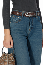 A close-up of a person wearing blue jeans with a brown leather belt featuring silver star details and a large silver buckle, paired with a dark gray ribbed top and holding a small woven handbag.