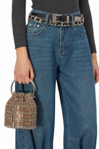 A person wearing blue wide-leg jeans, a black belt with a patterned strap, a dark gray ribbed long-sleeve top, and holding a small metallic rhinestone bucket bag against a plain white background.
