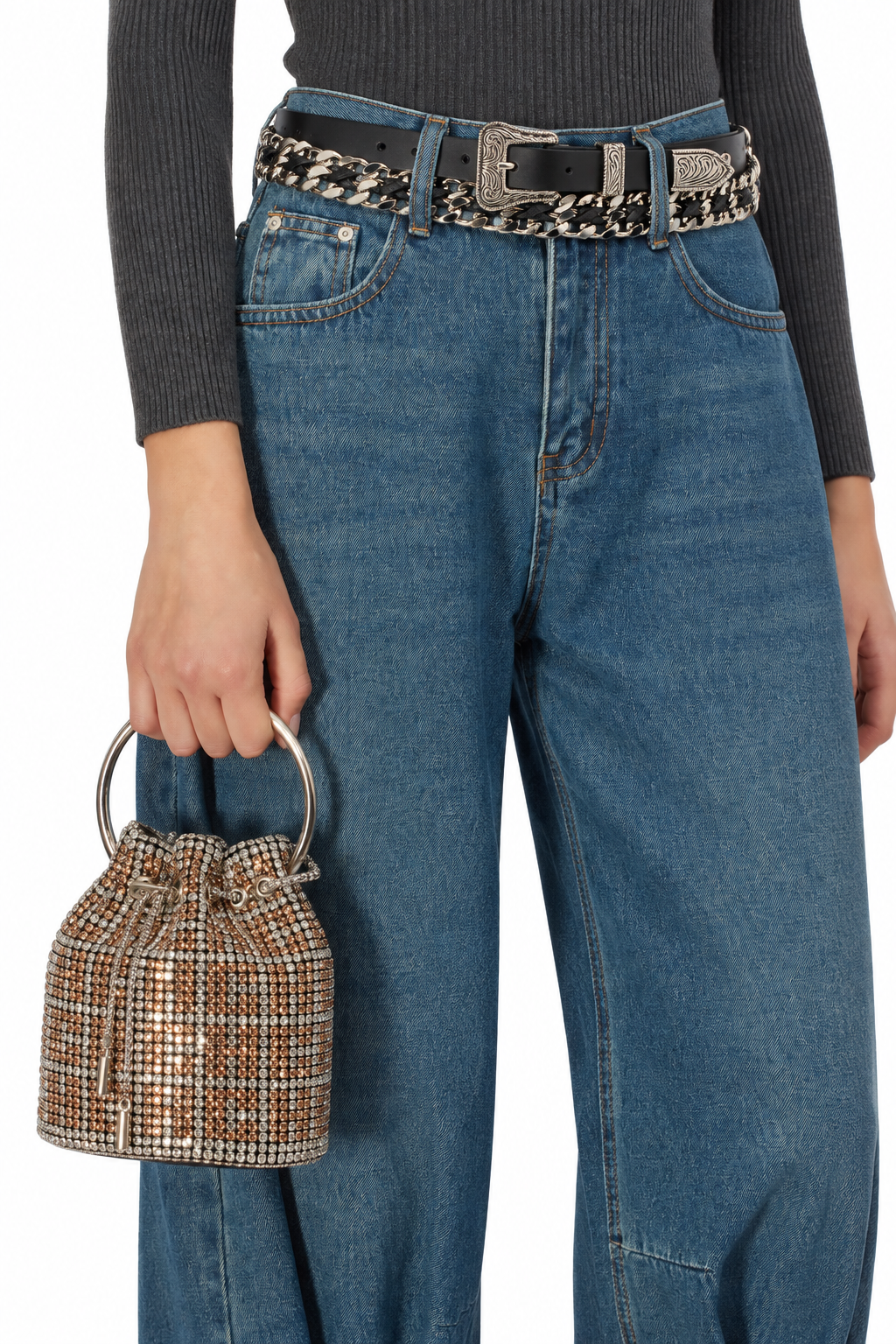 A person wearing blue wide-leg jeans, a black belt with a patterned strap, a dark gray ribbed long-sleeve top, and holding a small metallic rhinestone bucket bag against a plain white background.