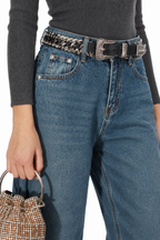 A close-up of a person wearing blue jeans with a black belt featuring a silver chain and ornate silver buckle, paired with a dark gray ribbed top and holding a small woven handbag.