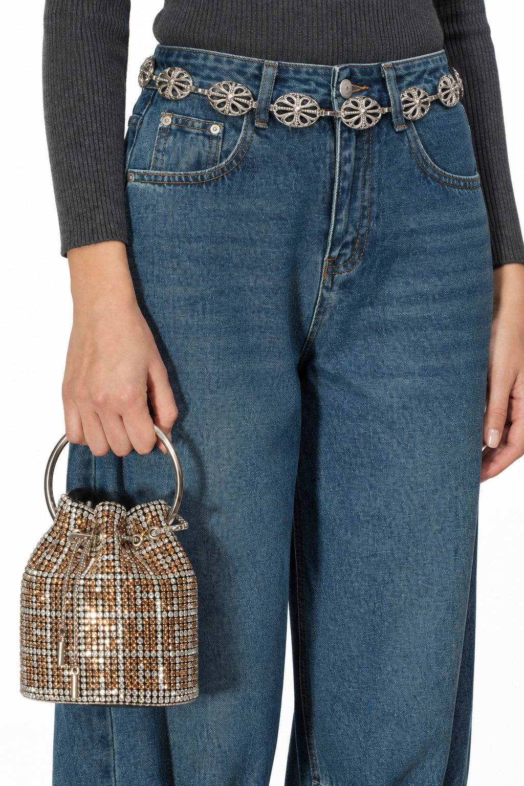 A person wearing blue jeans with ornate silver floral belt details, a dark gray ribbed top, and holding a small metallic gold and silver beaded bucket bag against a plain white background.