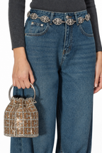 A person wearing blue jeans with ornate silver floral belt details, a dark gray ribbed top, and holding a small metallic gold and silver beaded bucket bag against a plain white background.
