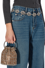 Close-up of a person wearing blue jeans with a decorative silver chain belt featuring circular medallion charms, paired with a dark ribbed top and holding a small rhinestone-embellished bucket bag.