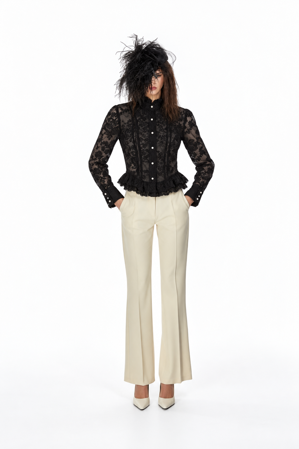 A model stands against a white studio background wearing a black lace long-sleeve button-front top with a ruffled hem, cream flared trousers, and a dramatic black feathered hat.