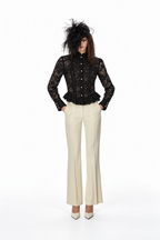 A model stands against a white studio background wearing a black lace long-sleeve button-front top with a ruffled hem, cream flared trousers, and a dramatic black feathered hat.
