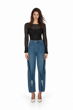 A model stands against a white studio background wearing a black long-sleeve lace top, blue wide-leg cropped jeans with silver zipper details at the ankles, and white pointed-toe heels.