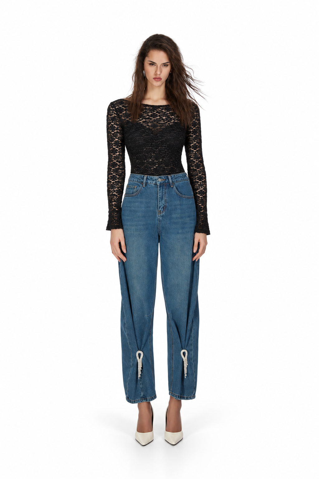A model stands against a white studio background wearing a black long-sleeve lace top, blue wide-leg cropped jeans with silver zipper details at the ankles, and white pointed-toe heels.