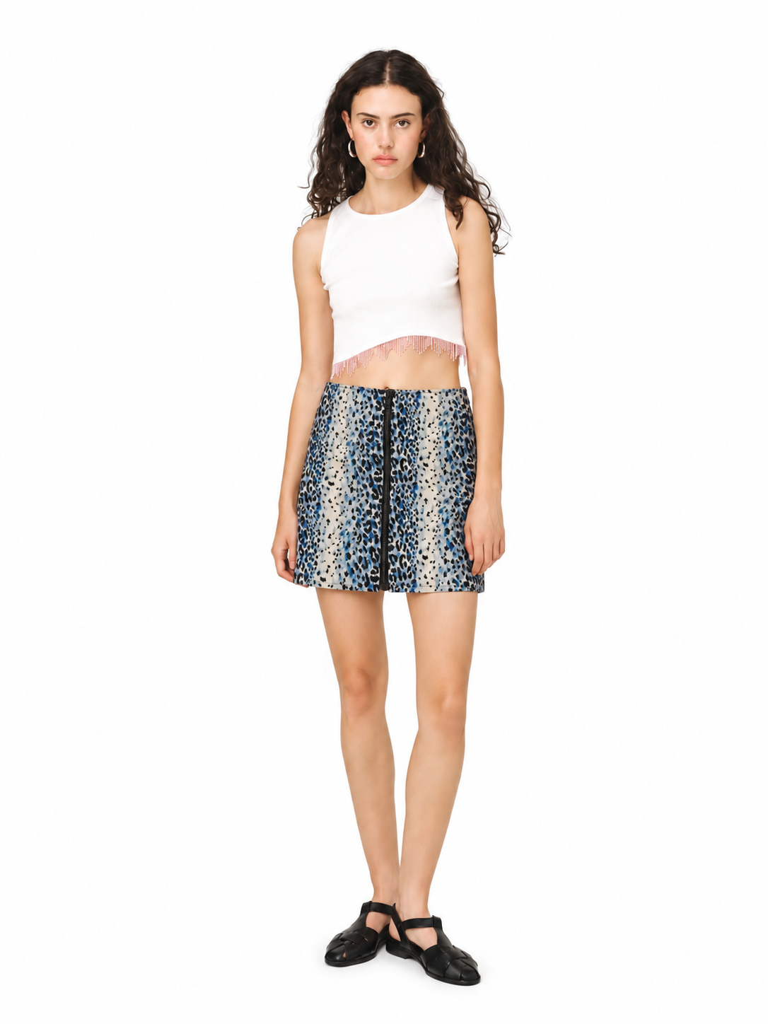 A model stands against a white studio background wearing a white sleeveless crop top with a pink ruffle hem, a blue and beige patterned mini skirt, and black Mary Jane shoes.