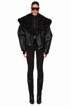 A model wearing a black faux-leather jacket with a large black fur collar, styled with black pants, black boots, and oversized sunglasses against a plain white background.