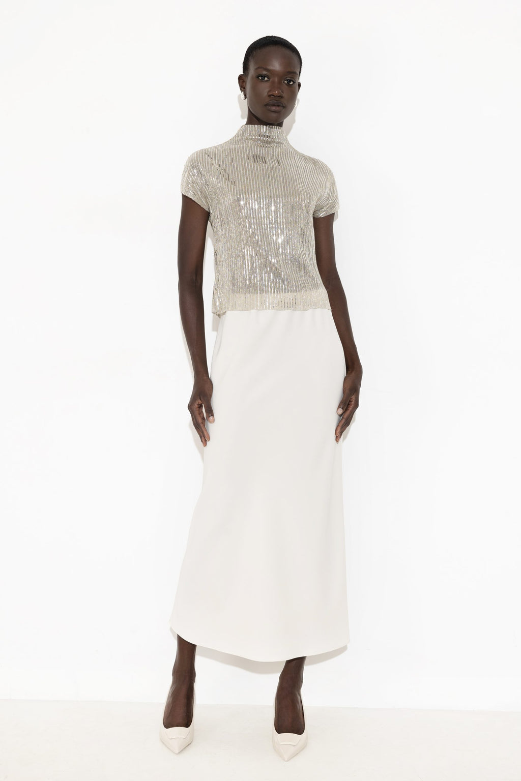 A model wearing a high-neck, short-sleeved dress featuring a shimmering silver sequined bodice and a solid white skirt, standing against a plain white studio background.