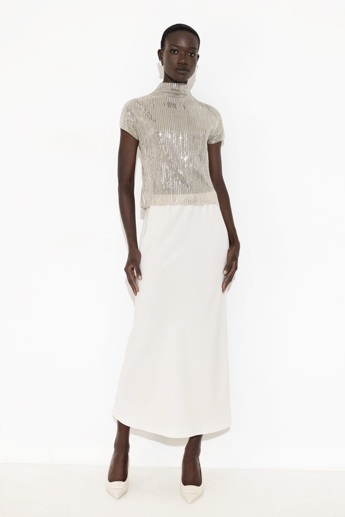 A model wearing a high-neck, short-sleeved dress featuring a shimmering silver sequined bodice and a solid white skirt, standing against a plain white studio background.