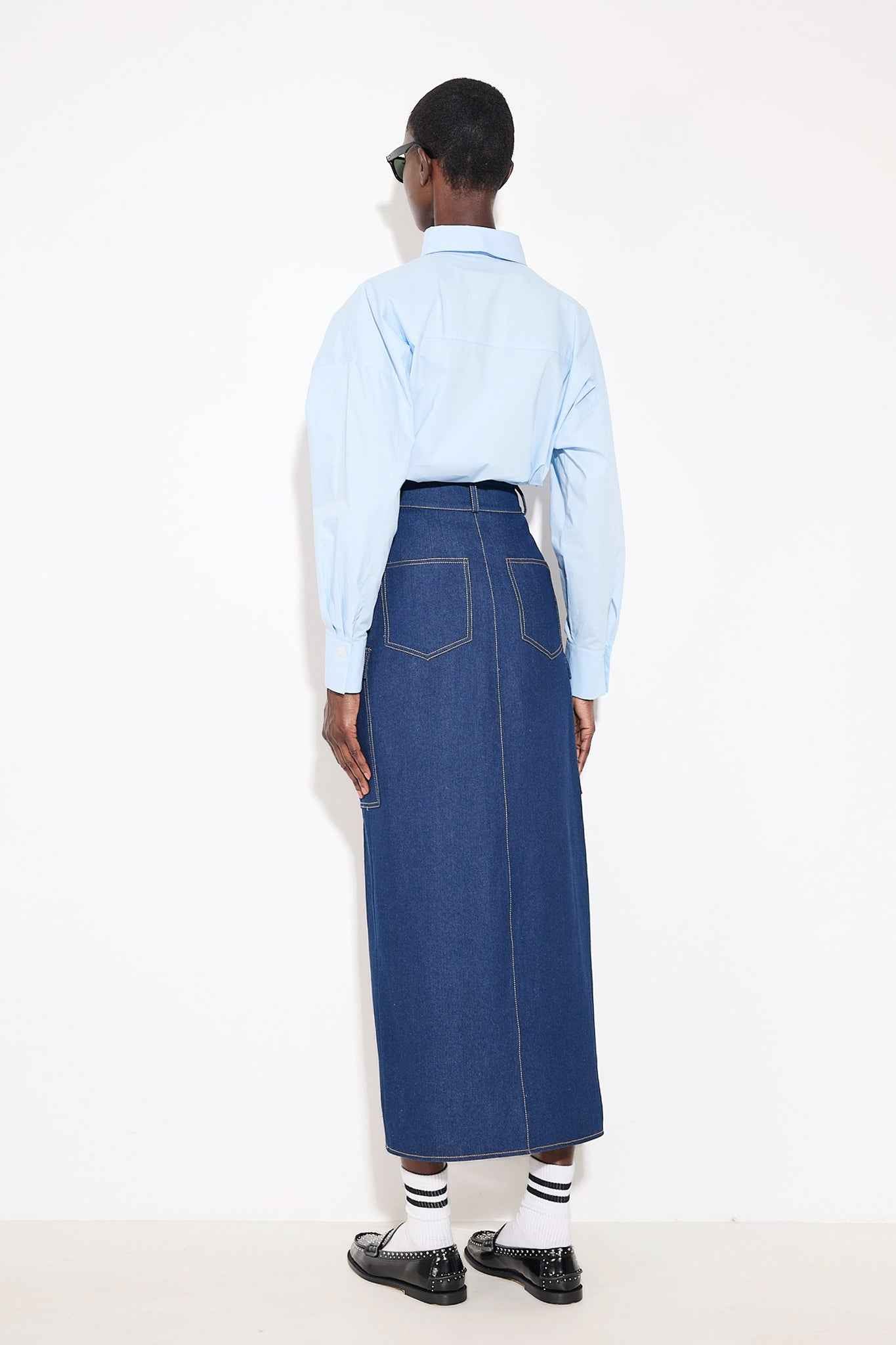 Person wearing a light blue shirt and long denim skirt on a white background