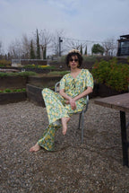 Woman in a floral dress sitting on a chair in an outdoor setting with plants and a table.