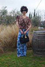 Woman in leopard print top and blue patterned pants holding a black handbag outdoors.