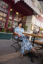 Woman in a blue floral outfit sitting on a chair outside a building with large windows.