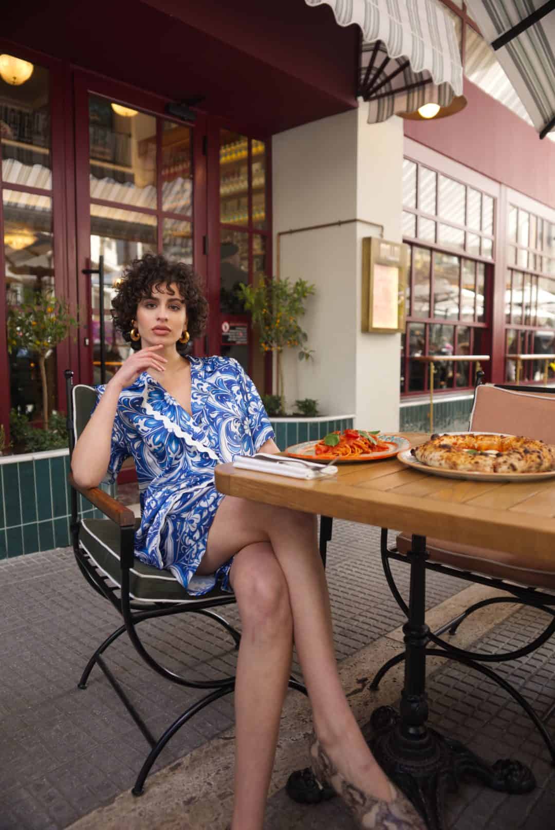 Woman in a blue and white dress sitting at an outdoor table with food.