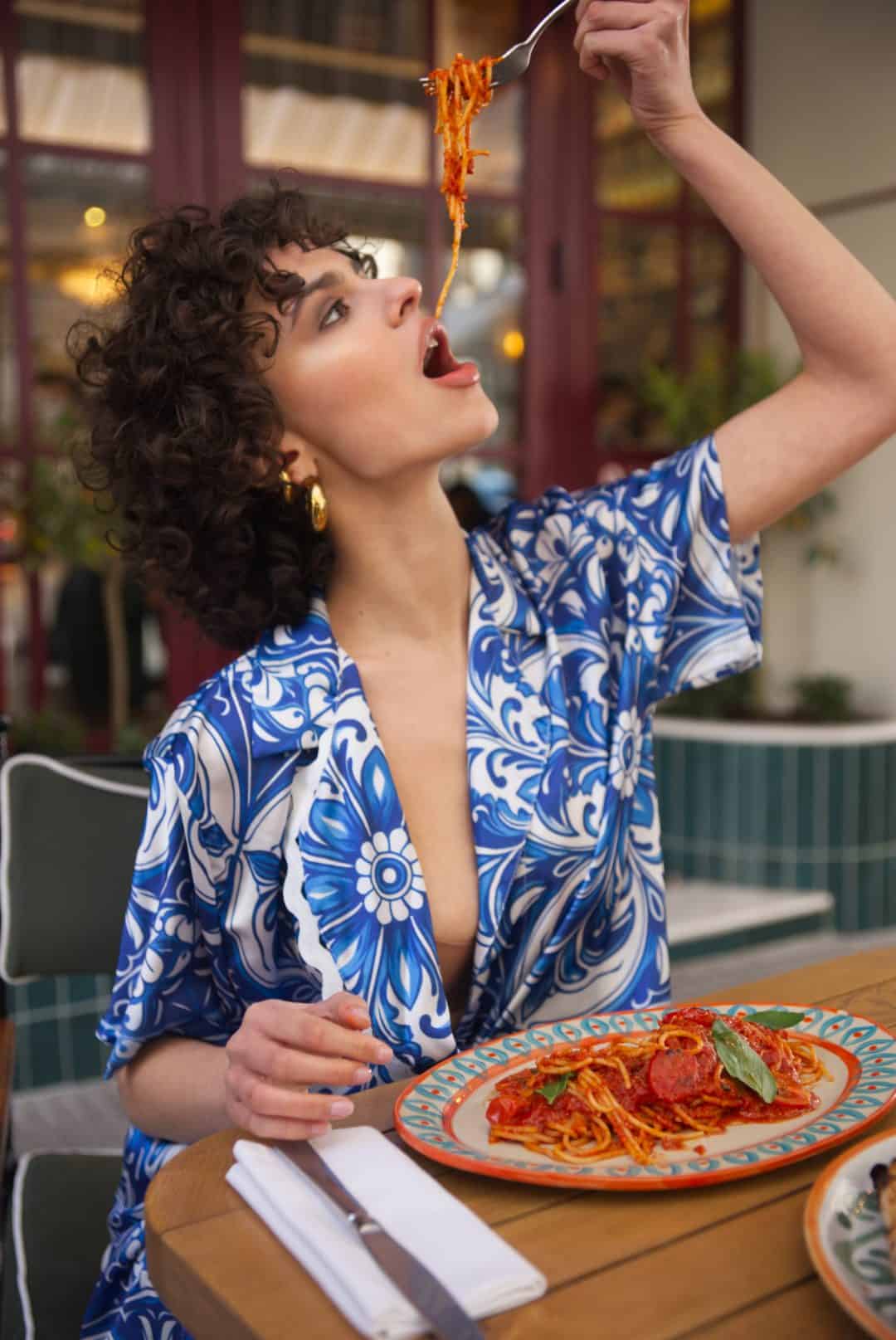 Woman in a blue and white patterned outfit eating spaghetti at an outdoor restaurant.