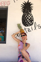 Woman holding a pineapple against a wall with a pineapple design and 'tails' text.